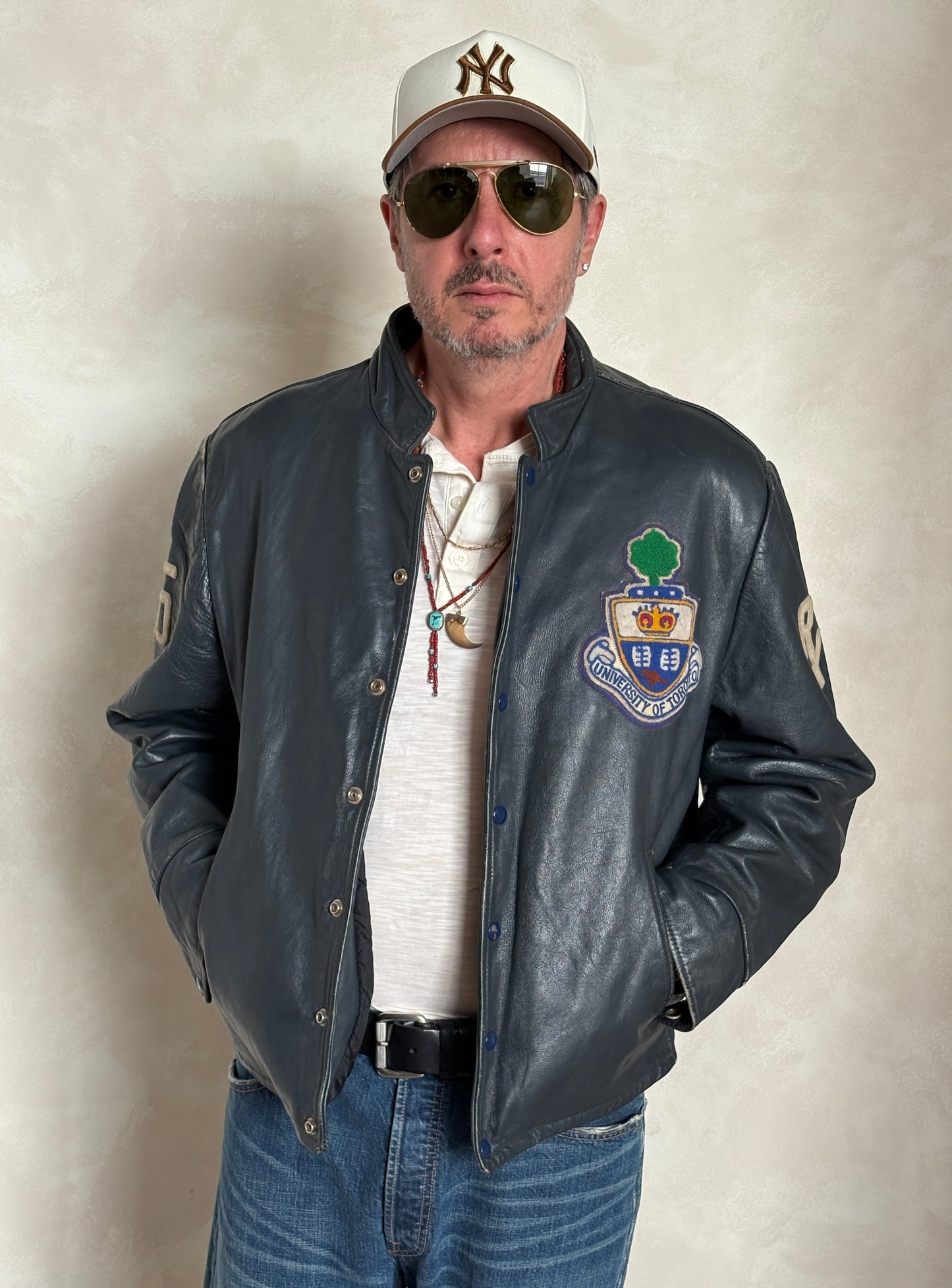 Man wearing a leather jacket with a logo, sunglasses, and a cap on a plain background