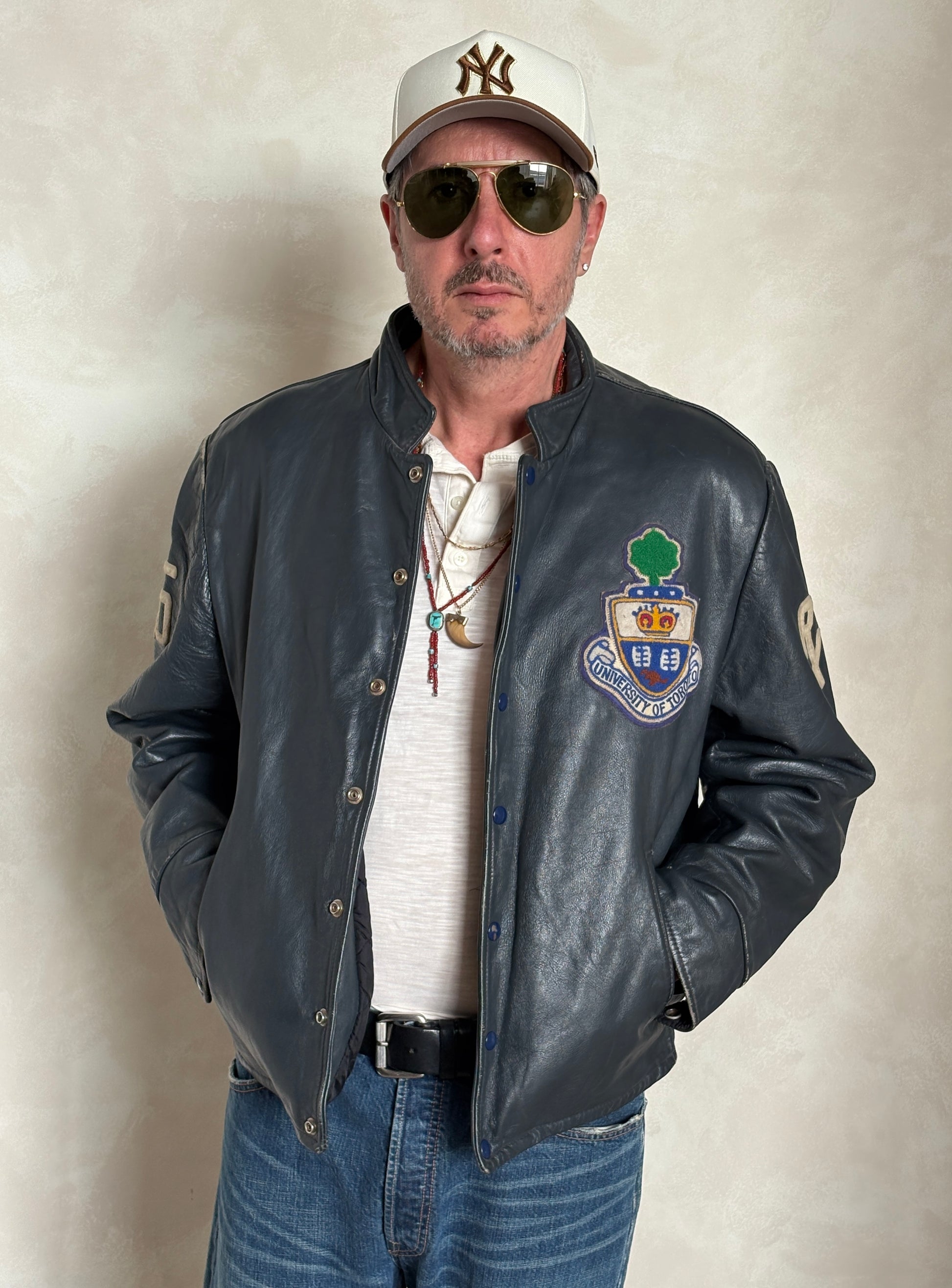 Man wearing a leather jacket with a logo, sunglasses, and a cap on a plain background