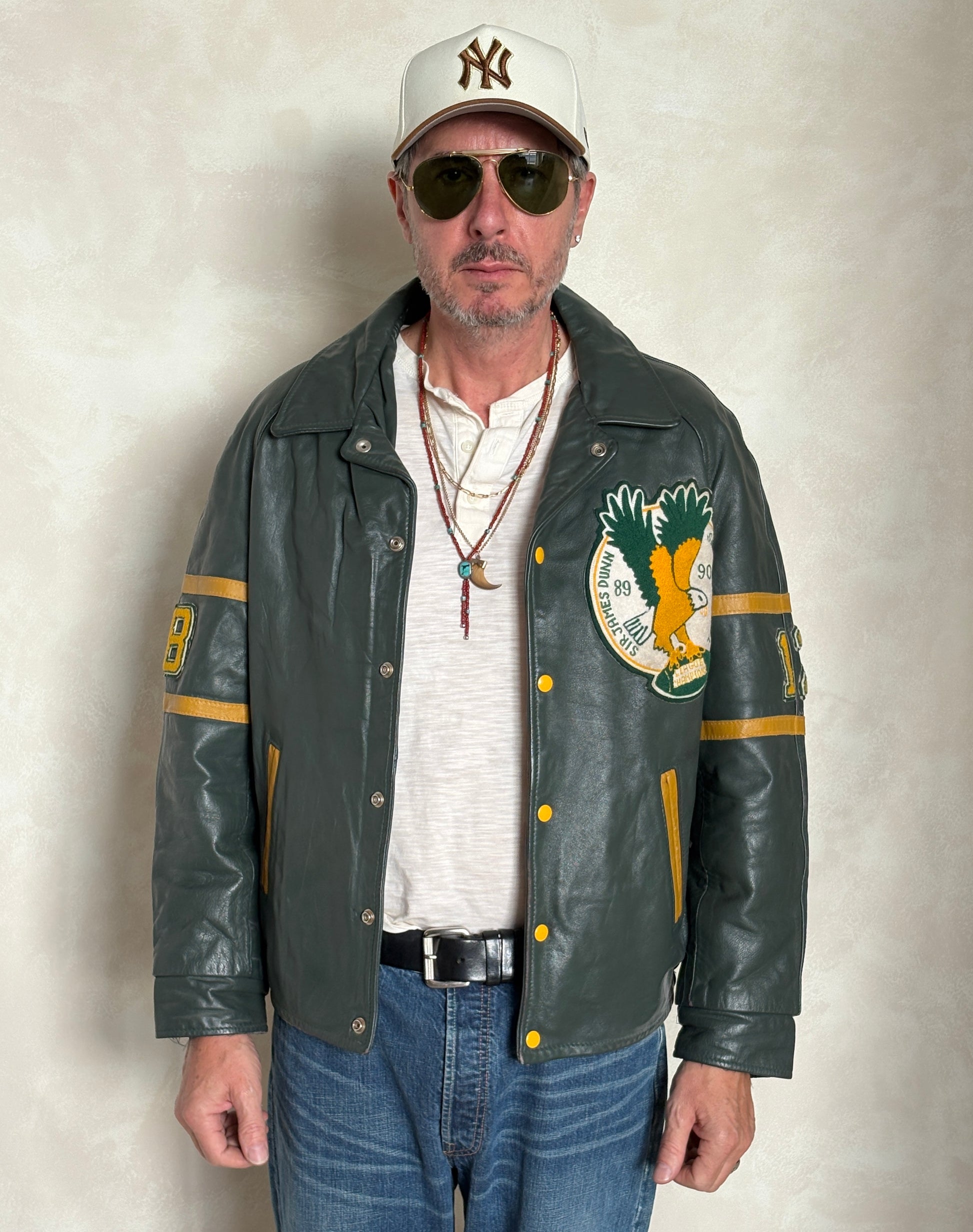 Man wearing a green leather jacket with patches, sunglasses, and a cap against a neutral background