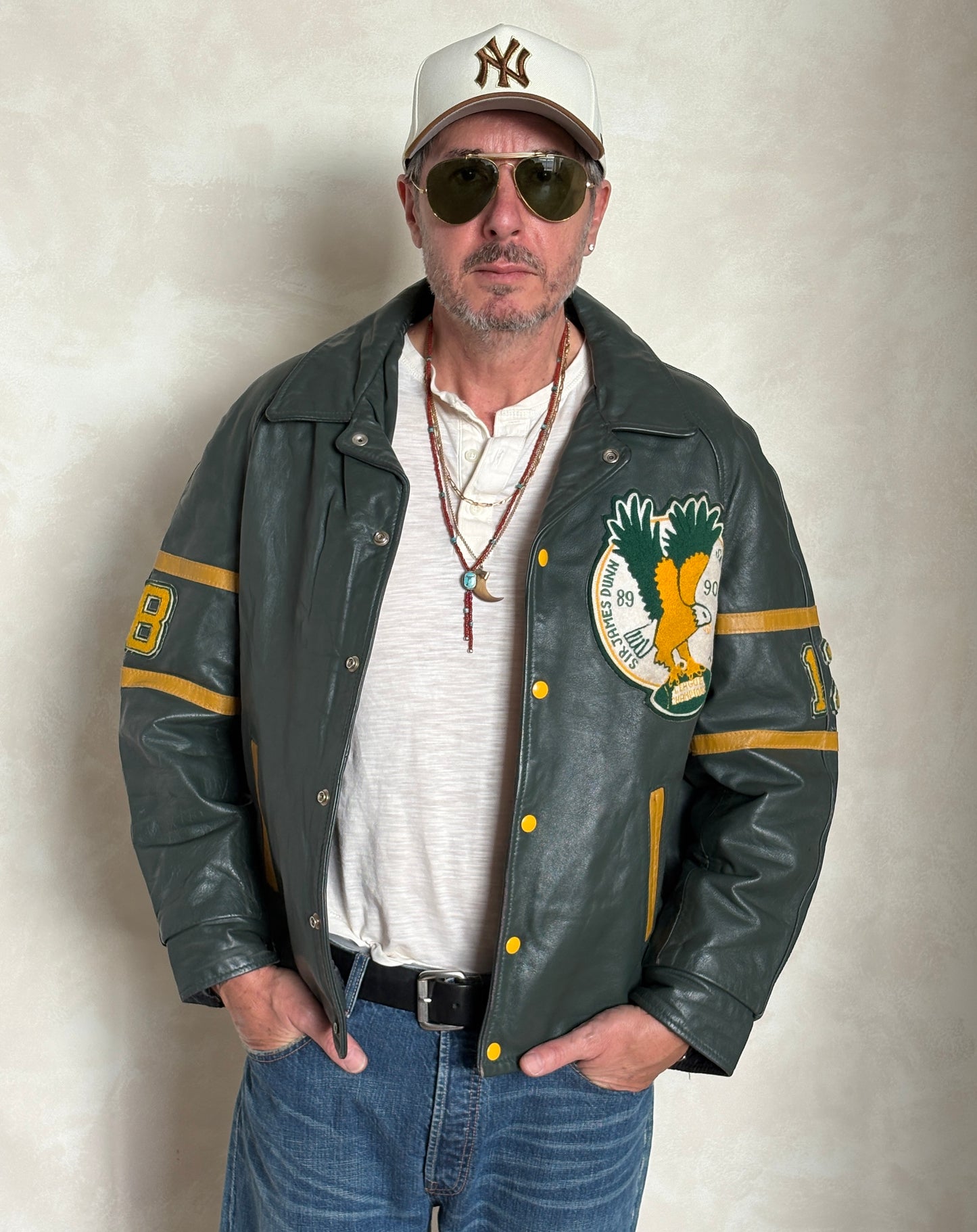 Man wearing a green leather jacket with a logo, sunglasses, and a cap against a plain background