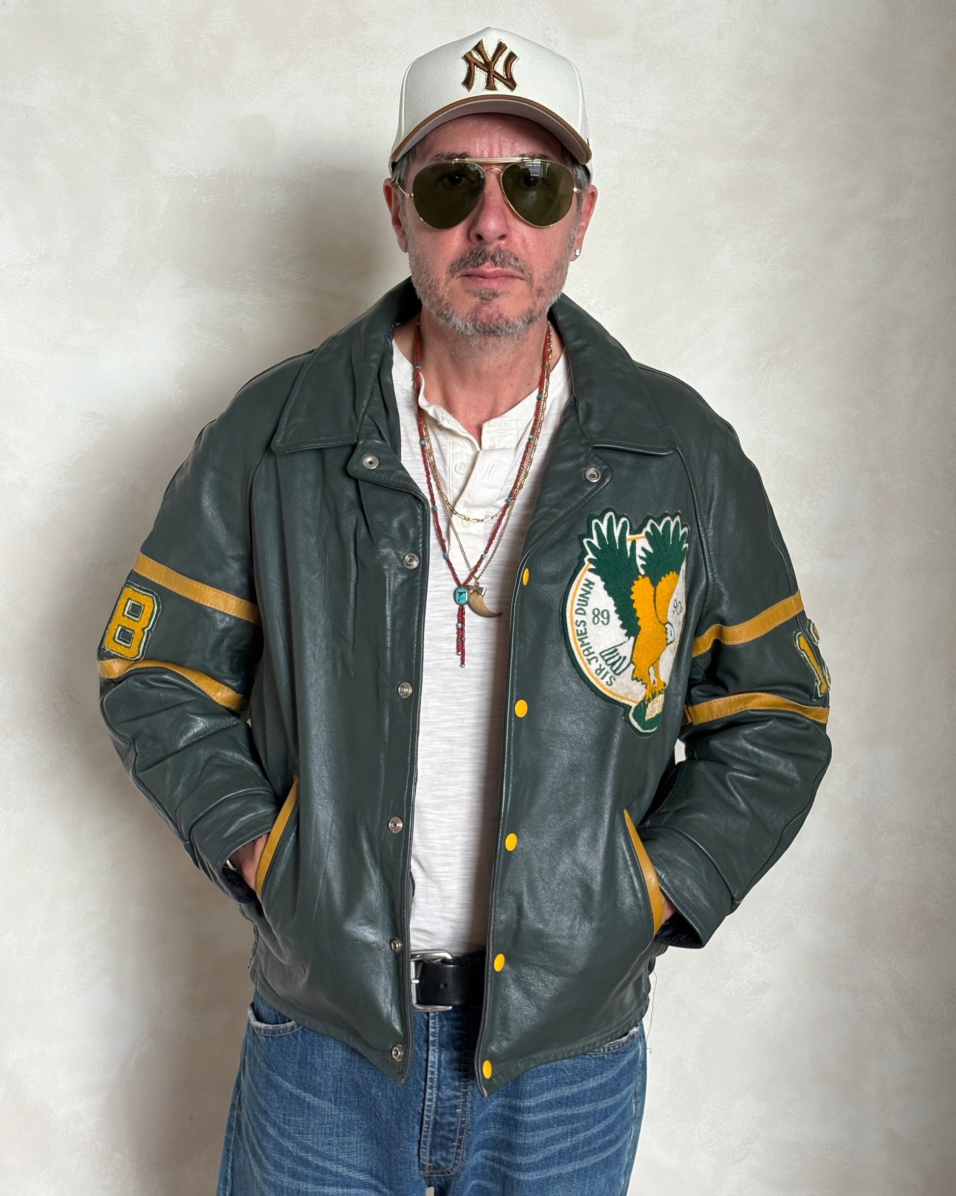 Man wearing a green varsity jacket with a logo, sunglasses, and a cap on a plain background