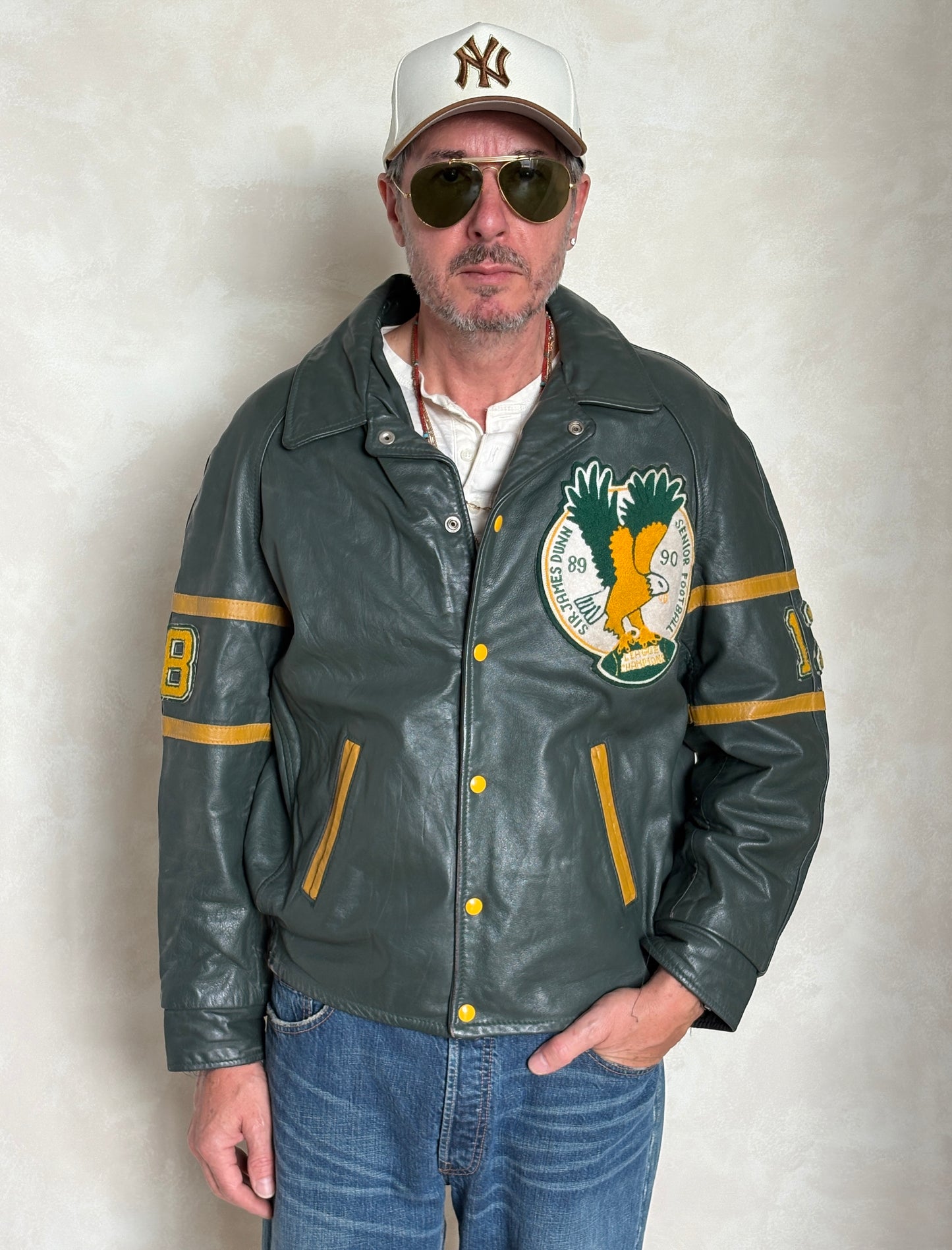 Man wearing a green leather jacket with a logo, sunglasses, and a cap on a plain background