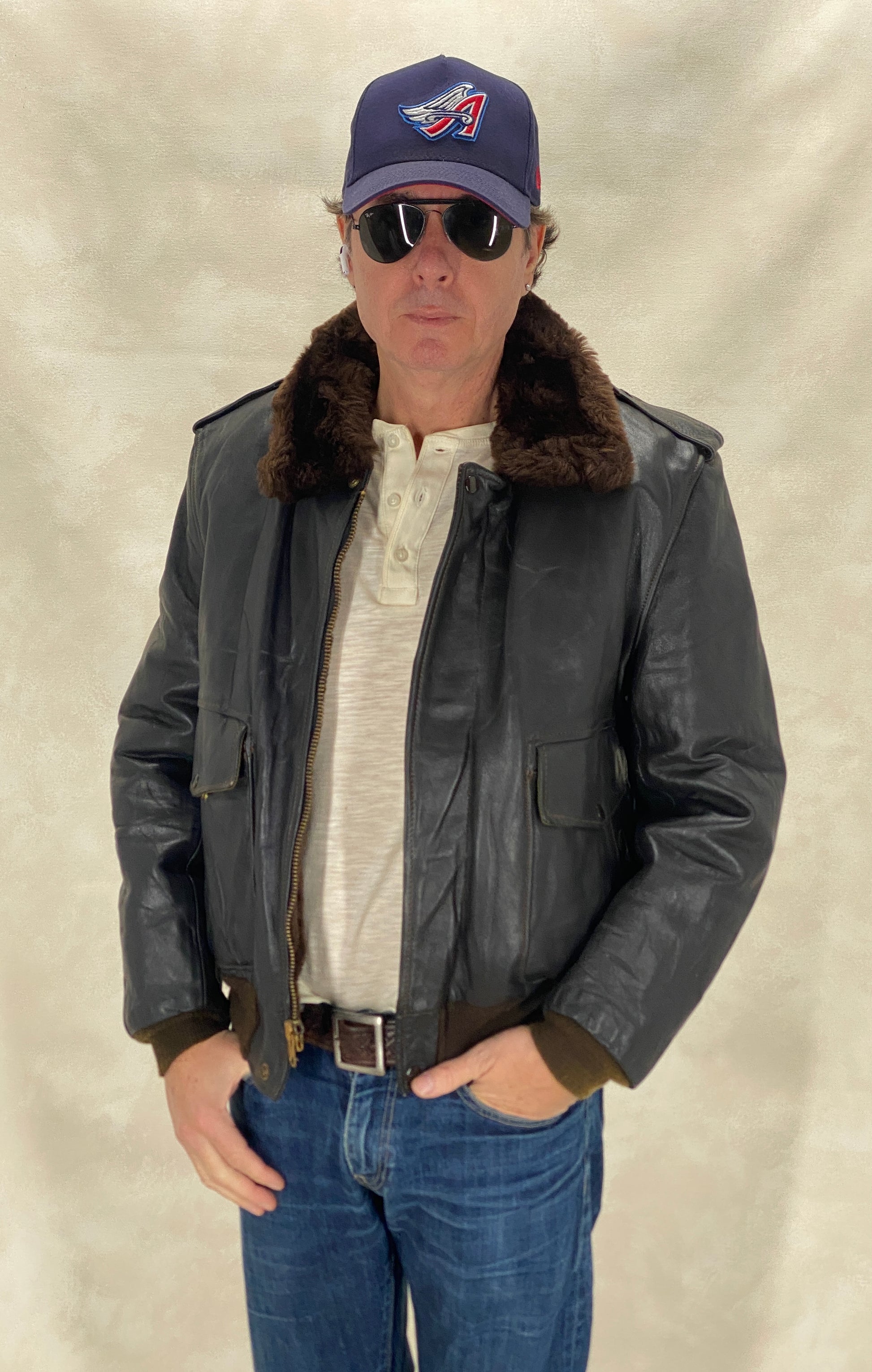 Man wearing a leather jacket with fur collar, sunglasses, and a baseball cap on a plain background