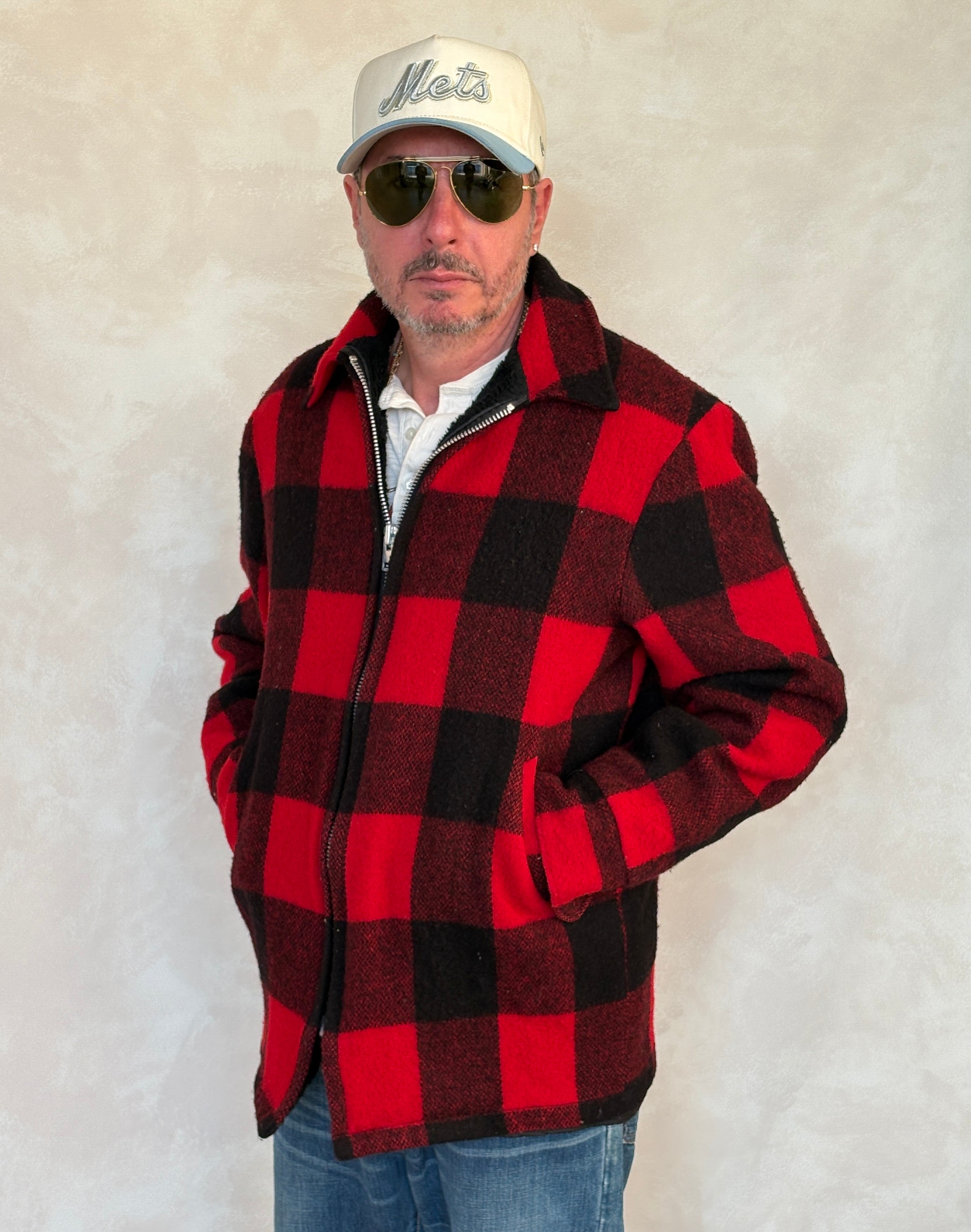 Man wearing a red and black checkered jacket, sunglasses, and a cap against a plain background