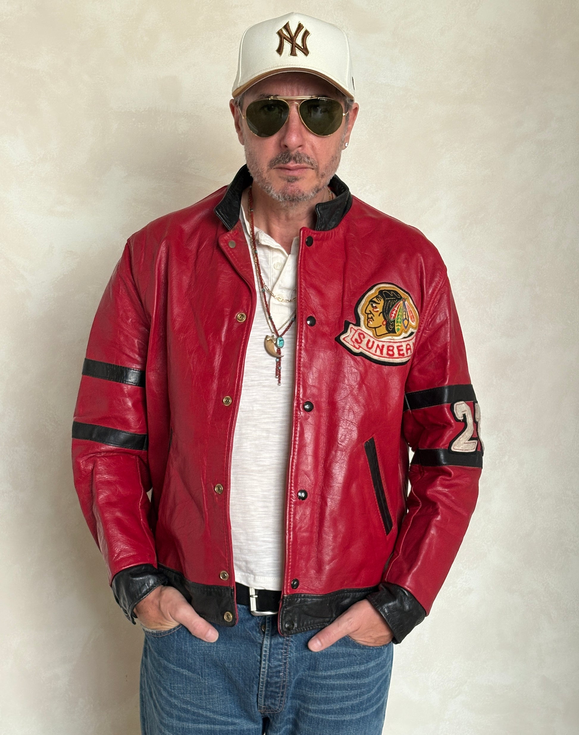 Man wearing a red leather jacket with patches, sunglasses, and a cap on a plain background