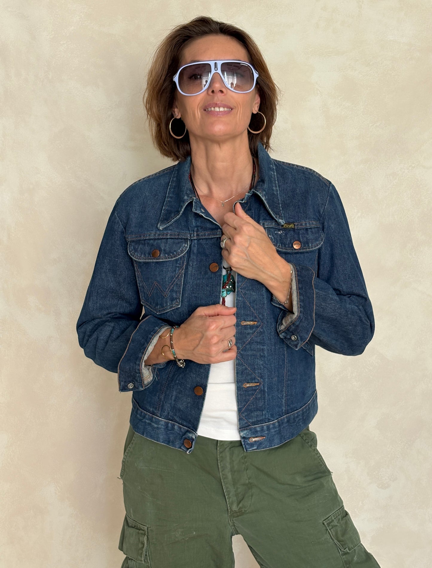 Woman wearing a denim jacket and green pants with sunglasses against a beige background